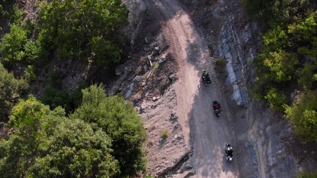 Aerial view of motorcyclists riding on a narrow rocky mountain road with sharp bends and steep cliffs. Adventure touring, remote travel, and rugged nature landscape captured in daylight.