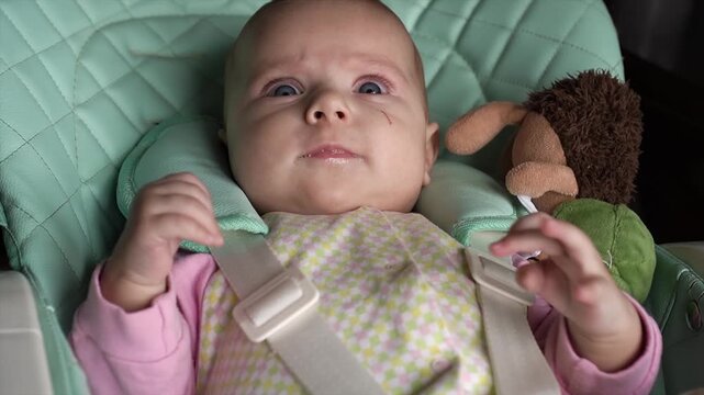 beautiful, happy, cute, surprised, excited little baby looks camera, with blue eyes, child's chair kitchen, children's clothes. smiles, amuses himself, shakes his legs, arms, drools. Dimples cheeks