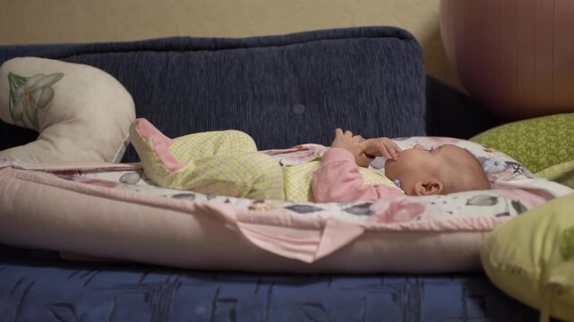 joyful little baby, lies calmly, puts his hands and fingers in his mouth, tastes, plays alone with himself, has fun, examines the room. A child in baby overalls, on a blue sofa in a cocoon. Side view