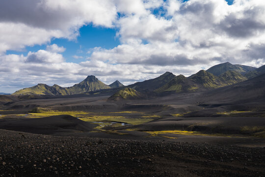 View of stark, dark volcanic landscape punctuated by patches of vibrant green moss and distant, craggy mountains under a sky of dramatic clouds, Skaftarhreppur, Iceland.