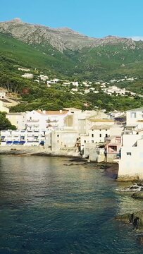 Charming coastal village with pastel buildings along the water, backed by lush green mountains in Erbalunga, Corsica. Bright, sunny day with clear blue sky.