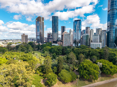 An aerial perspective of the lush Brisbane City Botanic Gardens contrasting with the modern central business district skyline under a bright, cloud-dotted sky.
