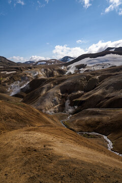 View of a landscape dominated by geothermal activity with steaming vents and snowy mountains under a bright blue sky in Hrunamannahreppur, Hrunamannahreppur, Iceland.