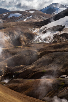 View of geothermal steam rises from the ochre and umber earth, contrasting with the distant snow-capped mountains under a dramatic sky, Hrunamannahreppur, Iceland.
