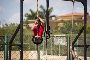 Obraz premium Middle Eastern man doing CrossFit rope climb with a friend in an outdoor fitness gym under the sun