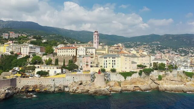 Aerial view of Bastia, Corsica, featuring pastel buildings along the rocky coastline with mountains in the background under a partly cloudy sky.