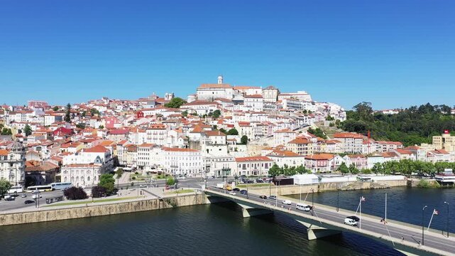Panoramic view of Coimbra s historic city center with red rooftops and the Mondego River under a clear blue sky. A bridge spans the river in the foreground, showcasing the vibrant architecture and