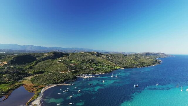 Stunning aerial view of Plage du Petit Sperone with turquoise waters, anchored boats, and lush green hills under a clear blue sky in Corsica.