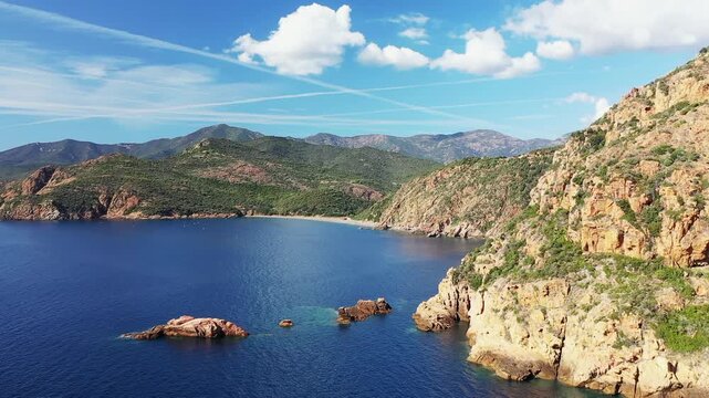 Scenic aerial view of the rugged coastline and clear blue waters of the Calanques de Piana in Corsica, France, with dramatic cliffs and lush green hills under a bright sky.