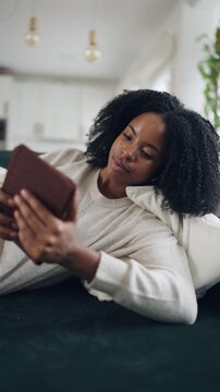Beautiful african american woman relaxing on the sofa at home while reading a book on her e reader, enjoying a peaceful domestic day