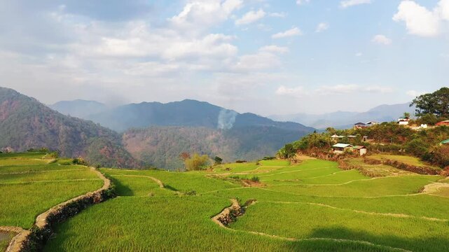 Lush green rice terraces stretch across rolling hills with distant mountains and scattered rural houses near Sagada, Philippines, under a partly cloudy sky.