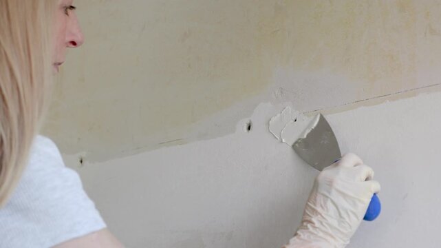 Woman with blonde hair in overalls fixing crack or hole in wall of apartment with plaster or putty and spatula tool to repair damage