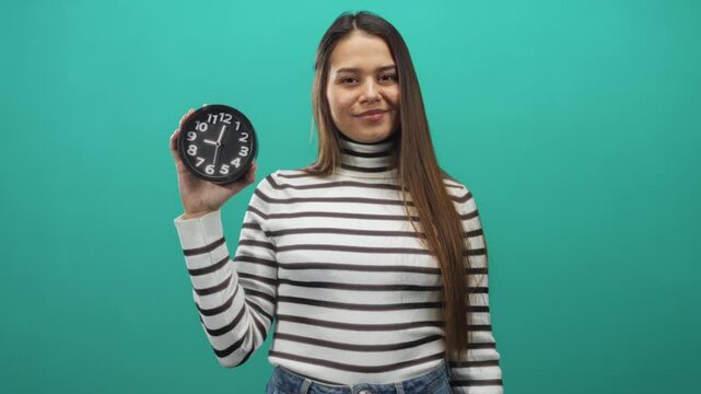 Woman holding black analog clock at chest level, smiling and showing thumbs up with other hand in studio green backdrop; optimism punctuality productivity.
