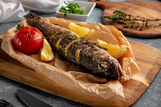 Roasted whole fish with crispy skin served on parchment paper with grilled tomato, lemon wedges and thyme on a rustic wooden board