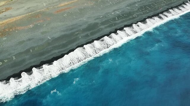 Aerial shot of waves crashing onto the unique black sand of Plage de Nonza in Corsica, with turquoise water and visible markings on the sand. The striking contrast between the beach and the sea