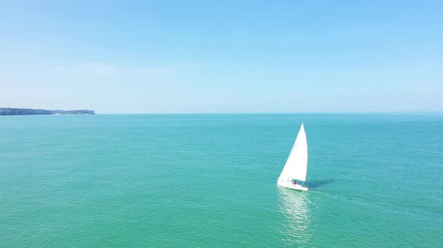 A white sailboat glides across the turquoise waters near the cliffs of Veules les Roses under a clear blue sky. Peaceful maritime scene with expansive open sea and distant coastline.