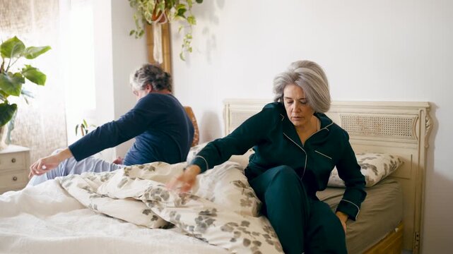 Elderly couple waking up together in a cozy bedroom. The senior woman stretches her aching back while her husband gets out of bed