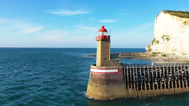 Aerial view of a lighthouse at the entrance of Port de Fecamp with dramatic white cliffs and coastal houses in Normandy, France. Clear blue sky and calm sea create a scenic maritime landscape.