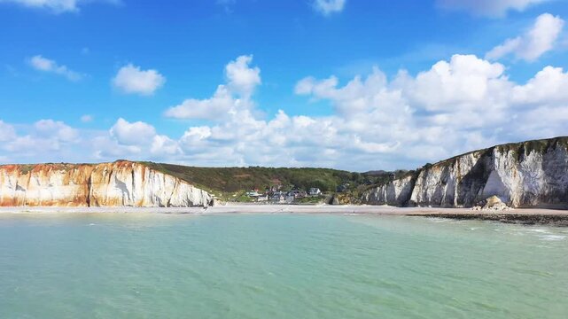 Scenic view of white chalk cliffs and a small coastal village at Les Petites Dalles under a bright blue sky with scattered clouds. Calm turquoise sea meets the dramatic coastline, creating a