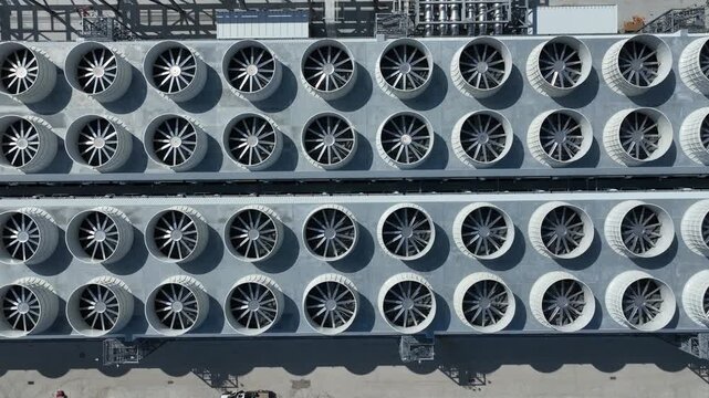 Aerial tilt from cooling fans to mechanical systems and data center buildings, revealing industrial infrastructure, piping, and HVAC supporting AI and cloud computing operations.
