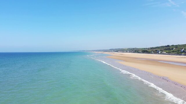 Wide view of Omaha Beach with gentle waves and golden sand under a bright blue sky. Tranquil seascape with distant shoreline and scattered houses.