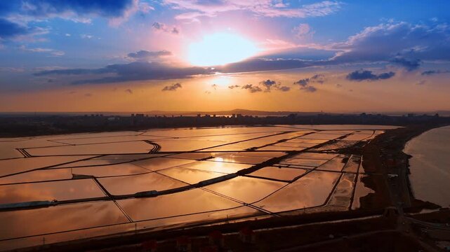 Extensive salt evaporation ponds reflecting vibrant sunset sky. Symmetrical grid of salt pans at dusk with sun reflection on water surface and distant city buildings.