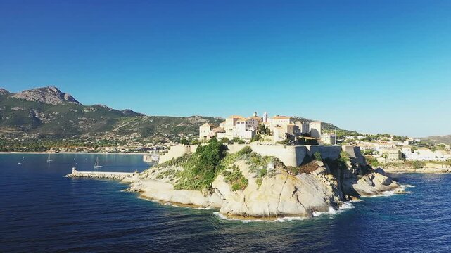 A scenic view of the historic citadel perched on a rocky promontory overlooking the blue Mediterranean Sea in Calvi, Corsica, with mountains and coastline in the background.