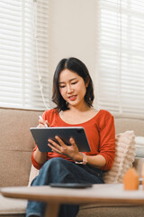 Asian woman using a digital tablet and stylus while relaxing on a sofa in a bright living room © PRIME STOCK LAB