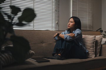 Naklejka premium Pensive young woman sitting alone on a sofa in a dimly lit living room with blinds