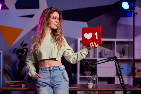 Young female blogger holding a like icon in a stylish workspace with colorful lighting, representing social media content creation