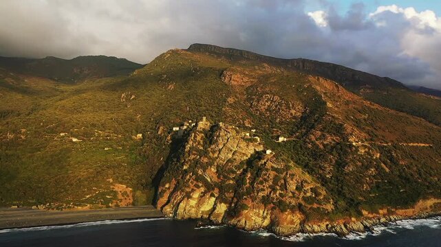 Aerial view of rugged cliffs and lush hillsides meeting the sea near Nonza, Corsica, under dramatic skies. The coastline features striking rock formations and natural Mediterranean beauty.