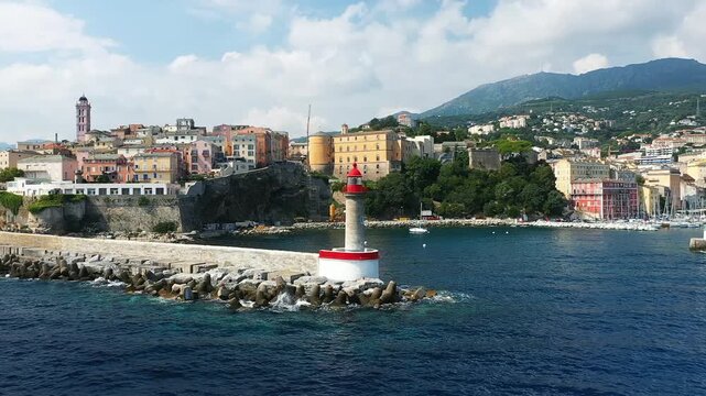 Scenic view of the Bastia waterfront with a red and white lighthouse, colorful historic buildings, and mountains in the background on the island of Corsica, France.