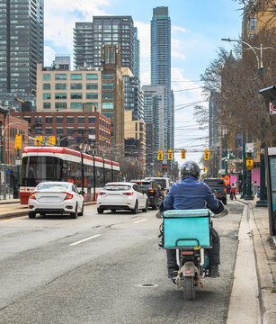 Delivery rider on moped with insulated cargo box navigating busy downtown street with mixed traffic and city skyline.