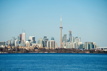 Obraz premium Panoramic view of the Toronto skyline featuring the CN Tower, seen from across Lake Ontario on a clear day. The cityscape showcases modern high-rise buildings and waterfront architecture with calm blu