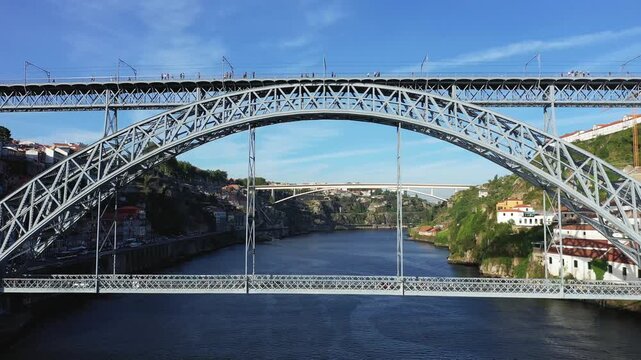 Scenic view of the Luis I Bridge spanning the Douro River in Porto, Portugal, with city buildings and hillsides under a clear blue sky.