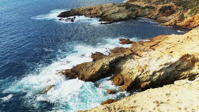 Aerial view of waves crashing against the rugged rocky coastline at Delta du Fango, Corsica. Clear blue sea contrasts with the textured golden rocks and sparse vegetation.