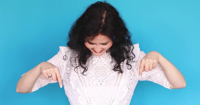 Young woman with curly hair and blue earrings in white lace dress pointing down with both index fingers on blue studio background, looking down at first then up at camera smiling, showing copy space