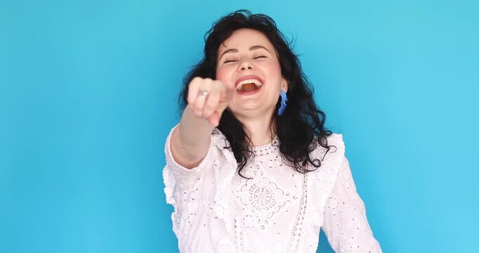 Energetic young woman with curly hair in white lace dress pointing finger at camera and laughing out loud, expressing great joy and amusement, throwing head back in laughter on blue background