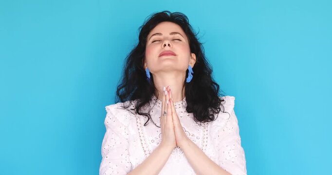 Young woman in white lace dress praying with hope, holding hands in namaste gesture with eyes closed then looking up with surprise and smiling on blue studio background