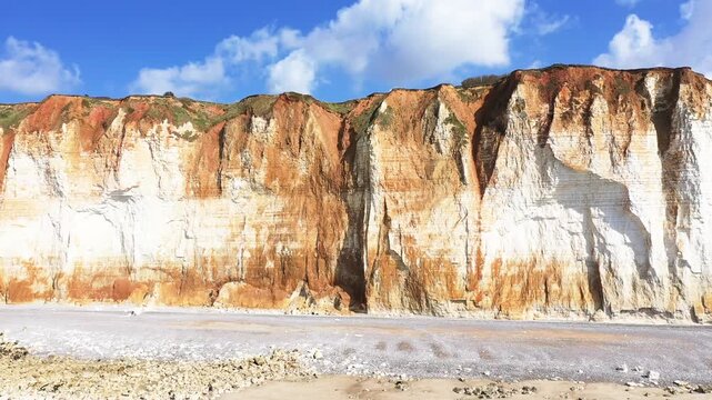 Dramatic white and ochre cliffs of Normandy rise against a bright blue sky with scattered clouds. Natural erosion patterns and vibrant colors highlight the rugged coastal landscape.