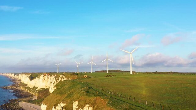 A row of wind turbines stands on green cliffs overlooking the sea in Fecamp, Normandy, France, under a clear blue sky. The dramatic coastline and renewable energy scene create a striking landscape.