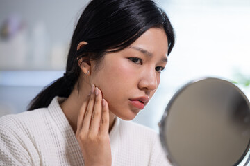 Young asian woman looking into round mirror touching face skin with hand showing worry about cheek...