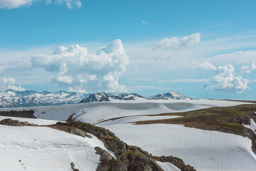 Scenic alpine landscape with sunlit snowfield with snow cornice above abyss against large snow-capped mountain range under cloudy sky. Snowy field near rocky precipice edge. High mountains in sunlight © Daniil