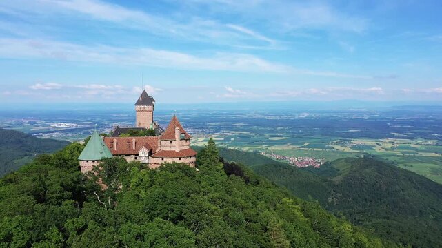 Aerial view of Chateau du Haut Koenigsbourg perched atop a lush green hill, overlooking the Alsace countryside under a clear blue sky.