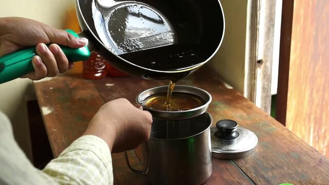 Close up of woman hands filtering used cooking oil into stainless steel container on wooden table, kitchen waste recycling concept