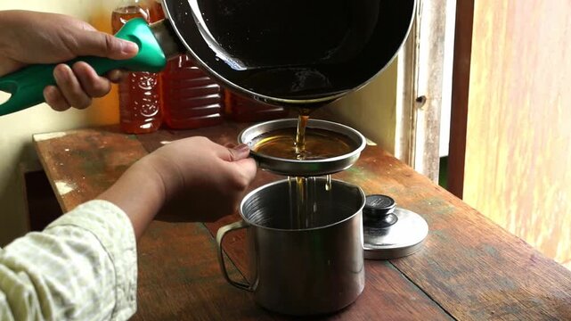 Close up of hands filtering used cooking oil from frying pan through strainer into stainless steel container, kitchen waste recycling and management concept