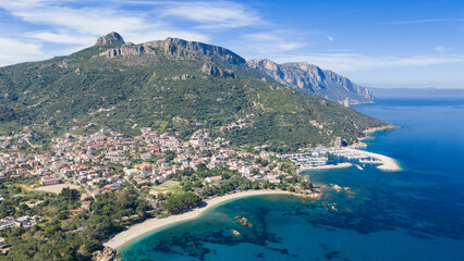 Obraz premium Aerial drone view of Santa Maria Navarrese with marina harbor, turquoise waters, sandy beach and dramatic limestone mountains along the eastern Sardinian coastline on a sunny day.