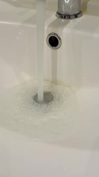 Dirty stainless faucet pouring water into unclean acrylic sink, visible mineral stains and soap scum around drain, slow steady.