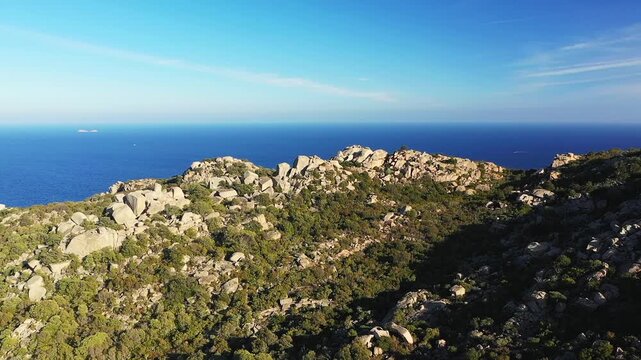 Aerial view of rocky hills covered with green vegetation overlooking the deep blue Mediterranean Sea near Plage de Santa Giulia, Corsica. Clear sky and natural landscape create a serene coastal scene.
