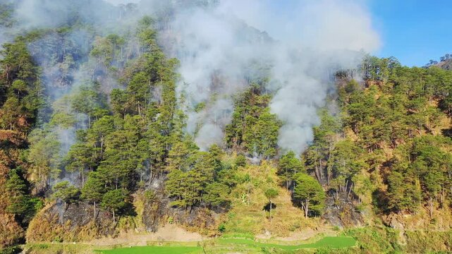 Dense smoke rises from a forest fire in the pine covered mountains near Bontoc and Sagada in the Philippines. The scene captures the impact of wildfire on the lush green landscape.